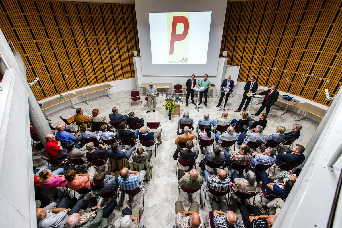 Leute sitzen in einem Saal vor einer grossen Leinwand.